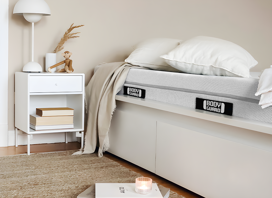 Een lichte kamer. Een BODYGUARD-matras ligt in een bed, daarnaast een nachtkastje met een houten aapje.