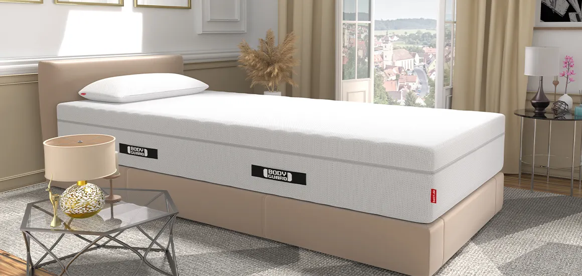 Die BODYGUARD Boxspring Matratze auf einem Bett in einem hellen Schlafzimmer. Auf der Matratze liegt das BODYGUARD Visco-Kissen. Im Hintergrund offenstehende, bodentiefe Fenster mit Blick auf eine Stadt, vorne ein Beistelltisch mit einer Lampe.